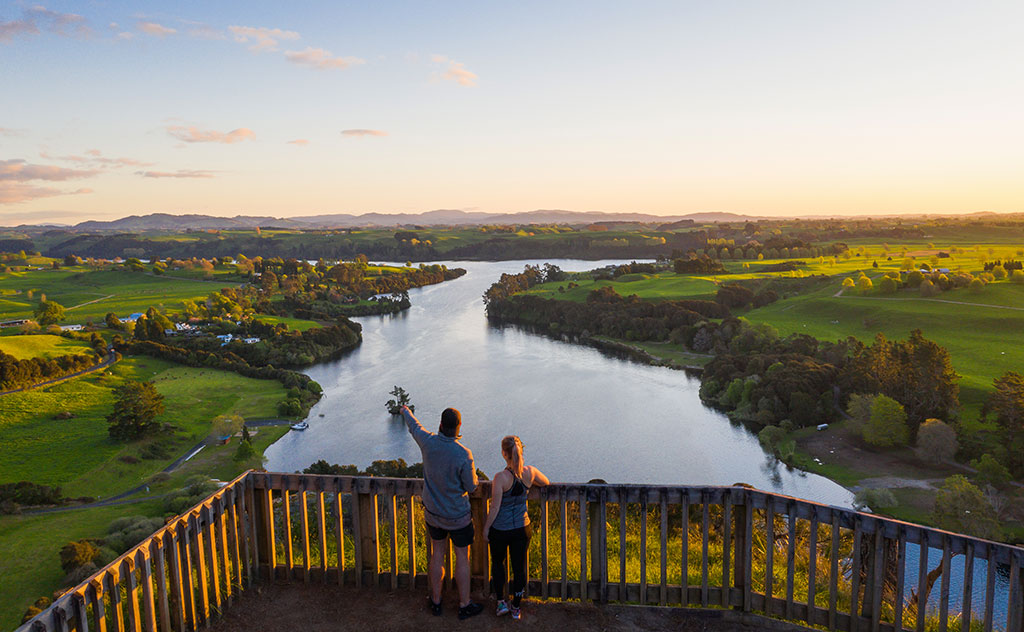 A man and a woman stand on a balcony overlooking the Waikato River as evening sun touches the riverbanks on either side of the river
