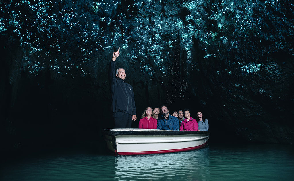 a man standing at the front of a small boat points to the roof of a cave illuminated by the glow of thousands of glow worms as guests watch on