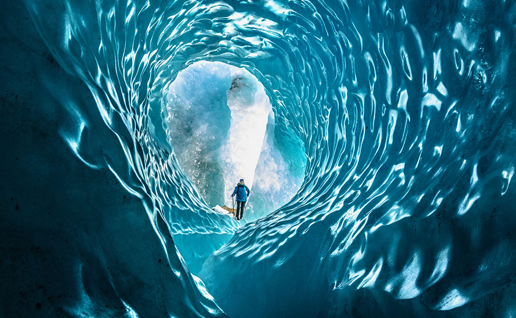 A tunnel of brilliant blue ice displays smooth pockmarks and leads to a sun-drenched glacier below at Tasman Glacier, New Zealand. A hiker stands at the bottom of the ice, admiring the view