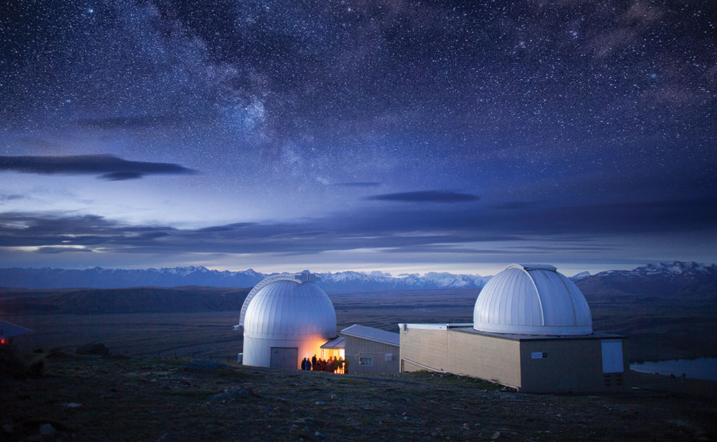 A blue evening sky is dotted with countless stars and delicate cloud formations above the Mount John Observatory near Tekapo, New Zealand. The Southern Alps are visible on the horizon, and a group of people stand outside one of the observatory buildings, cloaked in yellow light