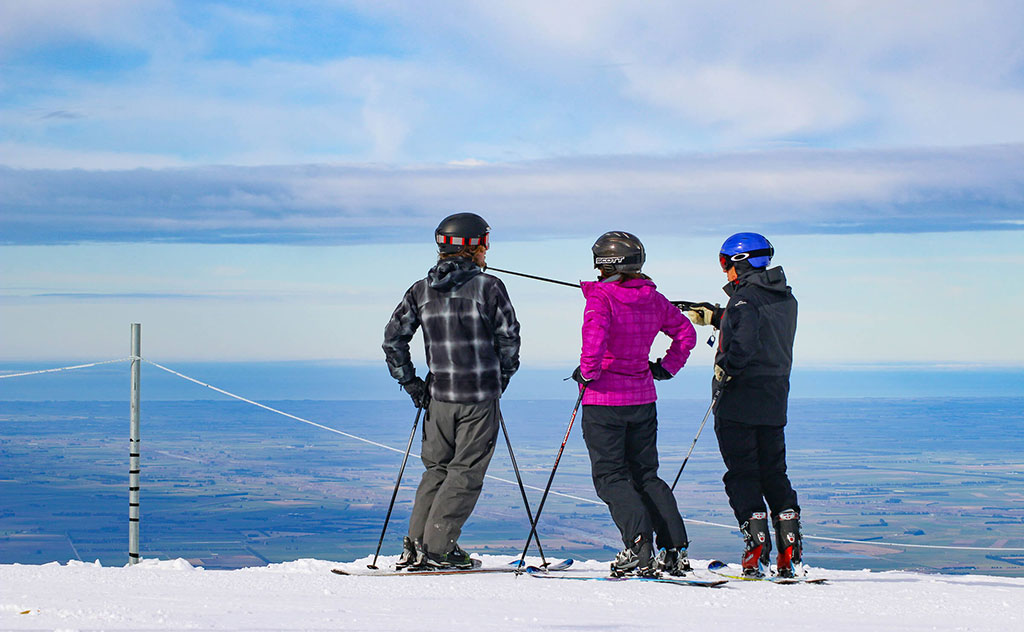 Three people wearing full ski gear with helmets and goggles stand on skis next to a boundary rope as they overlook the Canterbury Plains from Mt Hutt, New Zealand. One person uses a ski pole to point to an unseen feature in the distance