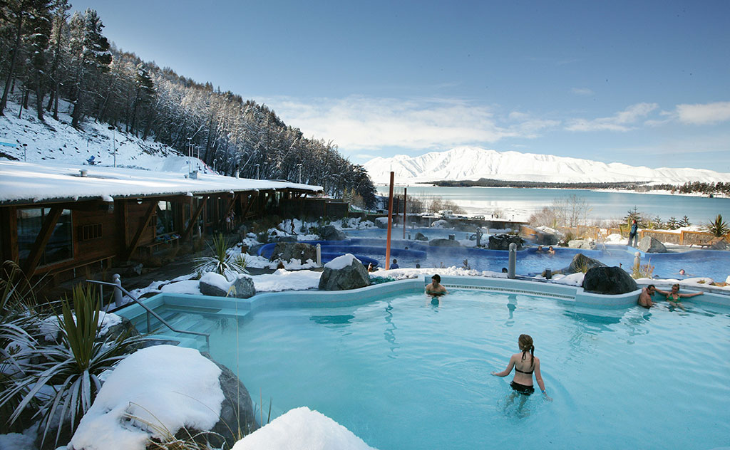 Tiered hot pools house guests in swimsuits at Tekapo Springs, New Zealand. Snow-capped hills can be seen in the distance, as steam from the hot water pools rises into the cold winter air