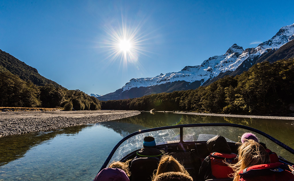 A jet boat filled with visitors navigates a shallow stretch of the Dart River, Queenstown on a clear-skied day, as the sun soars above snowy mountains