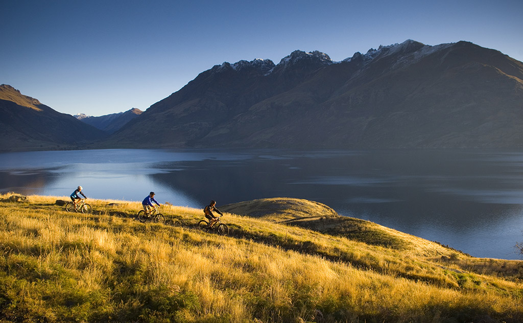 A trio of cyclists ride through tussock-lined banks on the shores of Lake Wakatipu as the afternoon sun sets over Jack’s Point, New Zealand