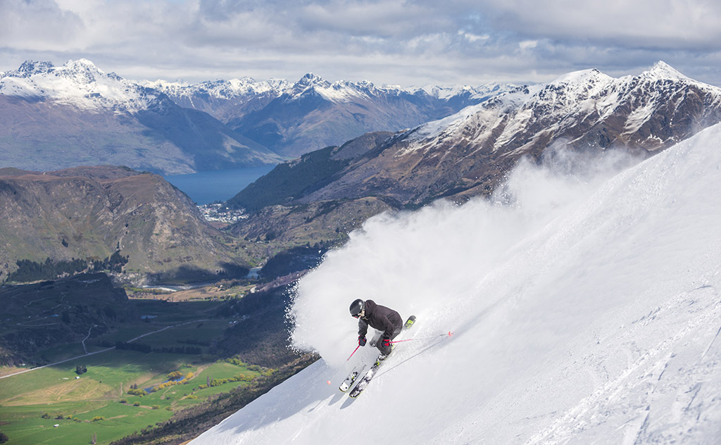 A skier wearing a black outfit and helmet makes a carving turn and leaves a high arc of snow spray behind on a cloudy day at Coronet Peak, New Zealand